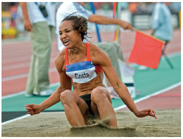 Imagem: Fotografia. Uma mulher com cabelos presos, usando top e short, com uma identificação no peito. Ela está com os pés sobre um quadrado com areia. Atrás, uma pista. Fim da imagem.