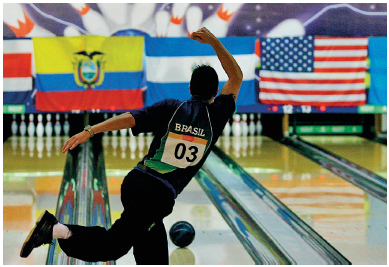 Imagem: Fotografia. Um homem de costas vestindo camiseta com ainscrição BRASIL está com uma perna e um braço levantado. À sua frente há uma pista de boliche com uma bola. Ao fundo,pinos de boliche. Fim da imagem.