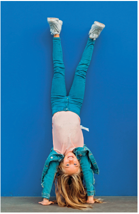 Imagem: Fotografia. Uma menina com cabelos loiros, usando camiseta, calça e tênis. Ela está com as mãos no chão e cabeça para baixo com as pernas estendidas para cima. Atrás, há uma parede. Fim da imagem.