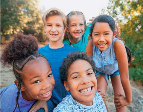 Imagem: Fotografia. Uma menina com cabelos crespos, usando camiseta azul, ao lado, um menino de cabelos crespos com camiseta estampada, atrás um menino com cabelos lisos, usando camiseta azul, ao lado, uma menina com cabelos lisos usando uma camiseta azul, ao lado, uma menina com cabelos pretos usando uma blusa regata. Eles estão juntos e sorrindo. Atrás, vegetação. Fim da imagem.