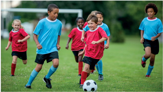 Imagem: Fotografia. Crianças com uniformes vermelhos e outros de azul correndo na direção de uma bola em um campo verde.  Fim da imagem.
