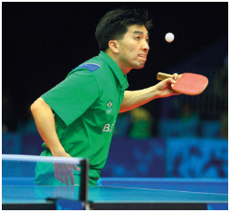 Imagem: Fotografia. Um homem usando uma camiseta verde, segurando uma raquete olhando na direção da bola, abaixo, uma mesa com rede. Fim da imagem.