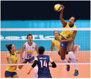 Imagem: Fotografia. Uma mulher uniformizada saltando com o braço esquerdo estendido na direção da bola, que está acima da rede. Atrás, outras mulheres uniformizadas olhando na direção da bola.  Fim da imagem.