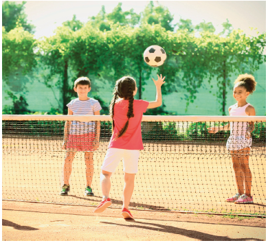 Imagem: Fotografia. Uma menina de tranças de costas com os braços estendidos na direção de uma bola. Atrás uma rede no chão e do outro lado, duas crianças olhando na direção da bola.   Fim da imagem.