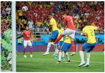Imagem: Fotografia. À esquerda, uma trave e um goleiro. Ao lado, uma bola no ar. Ao lado, jogadores com uniforme amarelo e vermelhos, saltando na direção da bola com as mãos próximas ao corpo  Fim da imagem.