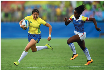 Imagem: Fotografia. À esquerda, uma mulher com uniforme verde e azul. Ela está correndo segurando uma bola de rugby em um campo verde. Ao lado, uma mulher negra usando roupa azul e branca, correndo na direção da mulher com a bola. Fim da imagem.