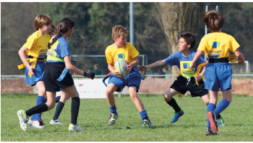 Imagem: Fotografia. À esquerda, um menino com uniforme azul e amarelo, ao lado, uma menina com uniforme azul e preto. No meio, um menino com uniforme azul e amarelo segurando uma bola. À direita, um menino com uniforme azul e preto com o braço estendido na direção do menino com a bola. Ao lado, outro menino com uniforme amarelo e azul de costas correndo na direção do menino com a bola.  Fim da imagem.