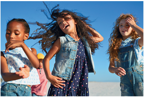 Imagem: Fotografia. Uma menina negra, com blusa estampada com as mãos na frente do corpo. Ao lado, uma menina com cabelos ondulados usando um vestido. Ela está com a mão esquerda na cintura e a mão direita na cabeça com os cabelos ao vento. Ao lado, uma menina ruiva, usando uma jardineira com a mão direita na cabeça.  Fim da imagem.