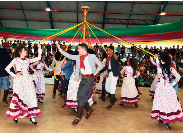 Imagem: Fotografia. Um grupo de homens com lenço no pescoço, calça e botas. Mulheres, com vestidos longos estampados e com babados e sapatos sociais. Eles estão em uma arena fazendo uma roda segurando uma fita presa a um pau no centro.  Fim da imagem.