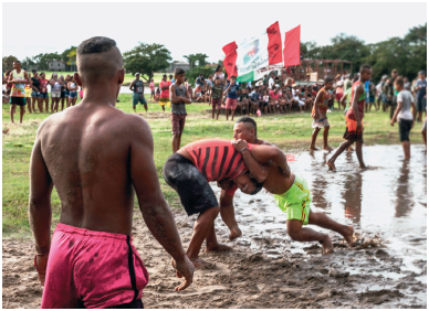 Imagem: Fotografia. Um homem com camiseta listrada e short. Ele está com as mãos ao redor dos braços de um homem com short amarelo no meio de um campo de areia e água. Ao redor, várias pessoas caminhando e outros parados olhando para eles.  Fim da imagem.