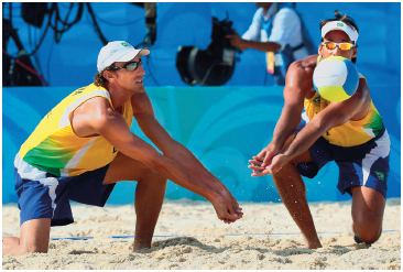 Imagem: Fotografia. Dois homens uniformizados agachados com os braços juntos na direção de uma bola em um campo de areia.  Fim da imagem.