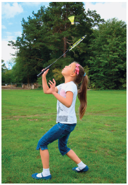 Imagem: Fotografia. Uma menina com cabelos loiros, usando uma regata e uma calça. Ela está de perfil segurando uma raquete na direção de uma peteca. Fim da imagem.