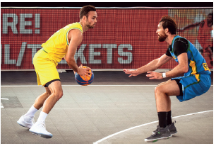 Imagem: Fotografia. À esquerda, um homem com uniforme amarelo, segurando com as mãos uma bola de basquete. Ao lado, um homem com uniforme azul. Ele está olhando para o homem com a bola com os braços estendidos para frente.  Fim da imagem.