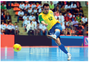 Imagem: Fotografia. Um homem com uniforme amarelo e azul, com a perna estendida na direção de uma bola em uma quadra. Atrás, uma arquibancada com várias pessoas olhando para ele.  Fim da imagem.