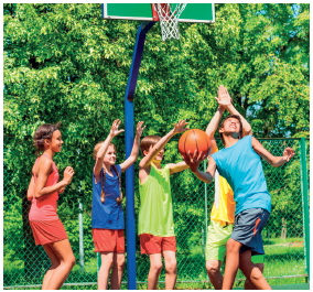 Imagem: Fotografia. Um grupo de crianças em uma quadra com cesta de basquete, com uma delas segurando uma bola de basquete. Fim da imagem.