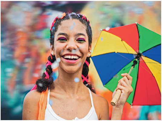 Imagem: Fotografia. Uma menina com cabelos presos com fita, segurando com a mão esquerda um guarda-chuva. Ela sorri. Confete coloridos caem do céu.  Fim da imagem.