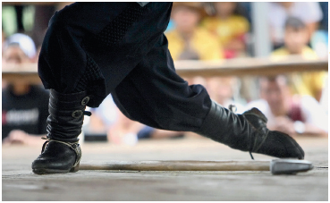 Imagem: Fotografia. Destaque de uma pessoa usando uma calça larga e uma bota. A perna direita está na frente e a perna esquerda estendida atrás e a direita.  Fim da imagem.