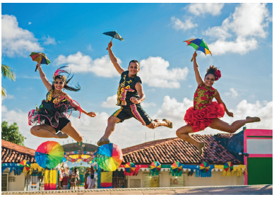 Imagem: Fotografia. Duas mulheres e um homem com roupas coloridas, segurando um guarda-chuva coloridos saltando com as pernas afastadas. Atrás, bandeirinhas coloridas.  Fim da imagem.