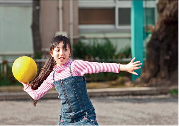 Imagem: Fotografia. Uma menina com cabelos lisos e presos, usando uma blusa listrada e uma jardineira. Ela está segurando uma bola com a mão esquerda.  Fim da imagem.