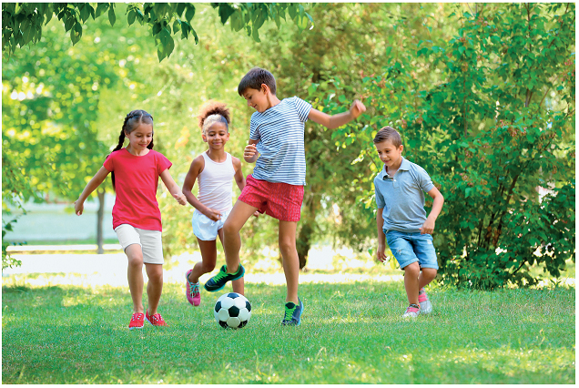 Imagem: Ilustração. Quatro crianças, dois meninos e duas meninas correndo atrás de uma bola na grama verde. Atrás, árvores.  Fim da imagem.