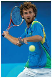 Imagem: Fotografia. Um homem com cabelos cacheados, usando uma camiseta azul, segurando uma raquete com a mão esquerda na direção de uma bola de tênis.  Fim da imagem.