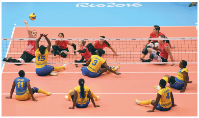 Imagem: Fotografia. Na frente, um grupo de mulheres com uniforme amarelo e azul, elas estão sentadas. Atrás, uma rede, e um grupo de mulheres com uniforme vermelho e preto, sentadas. Elas olham na direção da bola em cima da rede.  Fim da imagem.