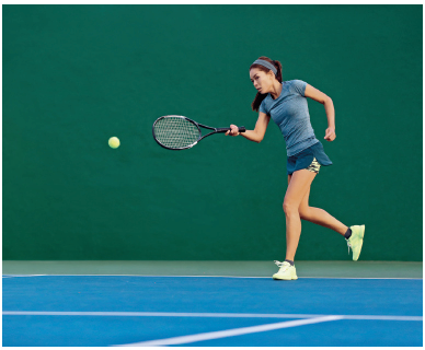 Imagem: Fotografia. Uma mulher com cabelos presos, usando camiseta cinza e saia. Ela está segurando uma raquete com a mão esquerda na direção de uma bola em um campo azul.  Fim da imagem.