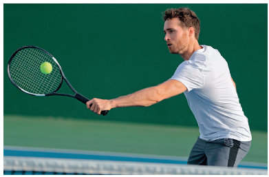 Imagem: Fotografia. Um homem com cabelos curtos, usando camiseta branca e short cinza. Ele está segurando uma raquete com a mão direita estendida para esquerda na direção de uma bola de tênis.  Fim da imagem.