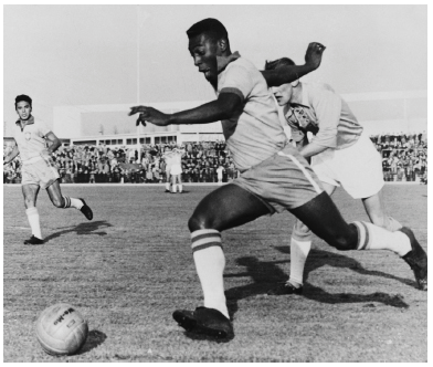 Imagem: Fotografia em preto e branco. Um homem negro com uniforme do Brasil Ele está correndo na direção de uma bola. Atrás, mais dois homens correndo no campo. Atrás, arquibancada com muitas pessoas.  Fim da imagem.