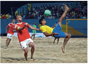 Imagem: Fotografia. À esquerda, um homem com uniforme vermelho e branco, ao lado, um homem com uniforme amarelo e azul. Ele está com a perna estendida no ar na direção de uma bola com as costas voltada para o chão de areia. No fundo, arquibancada, com pessoas sentadas.  Fim da imagem.