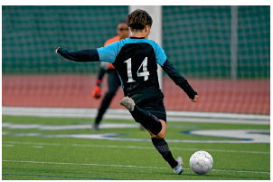 Imagem: Fotografia. Um menino com uniforme preto. Ele está de costas com a perna direita flexionada para trás na direção de uma bola. No fundo, um menino com uniforme vermelho e luvas. Atrás, o gol.  Fim da imagem.
