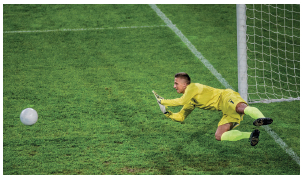 Imagem: Fotografia. Um homem com uniforme amarelo e luvas. Ele está deitado com o braço estendido na direção de uma bola. Atrás, a trave e o gol.  Fim da imagem.