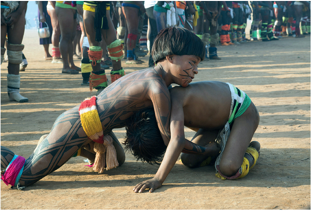 Imagem: Fotografia. Dois meninos indígenas com o corpo pintado. Eles estão de joelhos no chão agachado com os braços entrelaçados.  Fim da imagem.