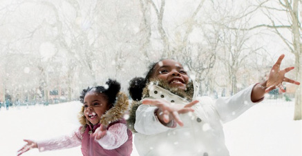 Imagem: Fotografia. Duas crianças com blusa estão sorrindo e com as mãos viradas para cima. Em volta delas há neve caindo e ao fundo, árvores cobertas de neve. Fim da imagem.