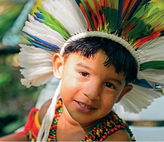 Imagem: Fotografia. Um menino indígena com cabelo curto, cocar colorido na cabeça e colar colorido no pescoço está sorrindo. Fim da imagem.