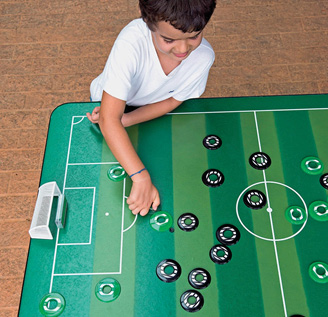 Imagem: Fotografia. Um menino está com a mão ao lado de um disco pequeno e verde, que está em cima de uma mesa com formato de campo de futebol. Em volta há discos verdes e pretos. Fim da imagem.