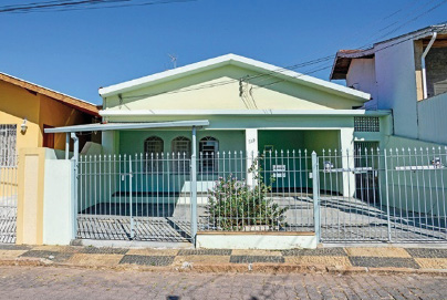 Imagem: Fotografia. Uma casa térrea com paredes verde-claro. Na frente há um portão de metal e nas laterais, mais casas.  Fim da imagem.