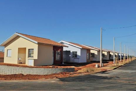 Imagem: Fotografia. Casas enfileiradas com paredes claras e telhado marrom. Ao lado há postes e uma rua asfaltada.  Fim da imagem.