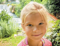 Imagem: Fotografia. Uma menina loira com olhos claros está sorrindo.  Fim da imagem.