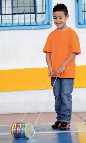 Imagem: Fotografia. Um menino com camiseta laranja, calça jeans e tênis preto está sorrindo e puxando duas cordas, que estão presas na lateral de uma lata colorida no chão. Fim da imagem.