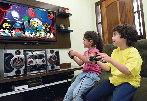 Imagem: Fotografia. Duas crianças estão sentadas em um sofá, sorrindo e segurando controles de videogame. Na frente delas há uma televisão com o jogo na tela.  Fim da imagem.