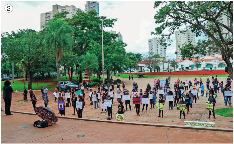 Imagem: Fotografia 2. Várias pessoas estão em uma praça, distantes umas das outras, com máscaras e segurando cartazes. Ao fundo, árvores e construções. Fim da imagem.