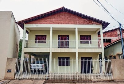 Imagem: Fotografia. Sobrado com paredes amarelo-claro e telhado marrom. Na frente há um portão de metal e nas laterais, mais casas.  Fim da imagem.