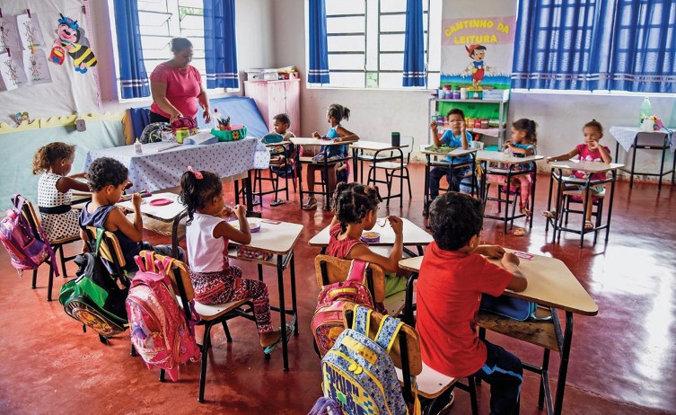 Imagem: Fotografia. Crianças estão sentadas em carteiras escolares e enfileiradas. Ao lado, uma professora está em pé, atrás de uma mesa com objetos em cima.  Fim da imagem.