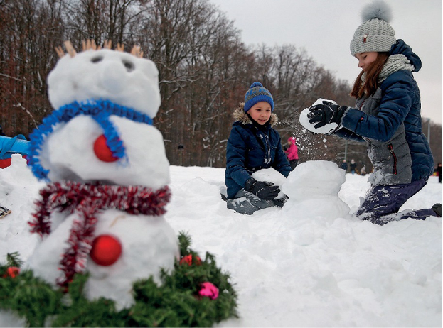 Imagem: Fotografia. Duas crianças com gorro, blusa, calça e botas estão ajoelhadas sobre a neve e montando um boneco. Ao lado, boneco de neve com cachecol azul e vermelho. Ao fundo, árvores secas. Fim da imagem.