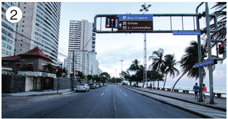 Imagem: Fotografia 2. No centro, uma rua asfaltada com um semáforo. À direita, árvores e o mar e à esquerda, casas e prédios. Fim da imagem.