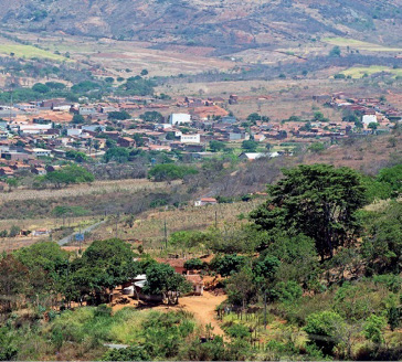 Imagem: Fotografia. Em primeiro plano há várias árvores. No centro, casas espalhadas e ao fundo, morros e árvores.  Fim da imagem.