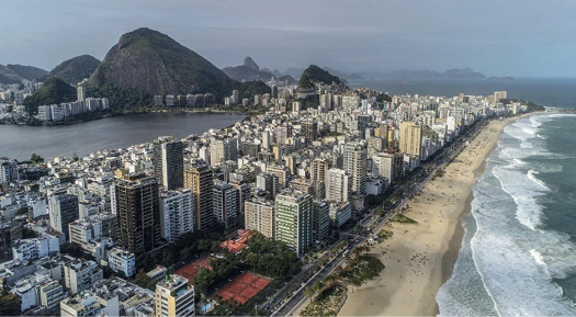 Imagem: Fotografia. À direita, a orla da praia. No centro, várias casas, prédios e árvores. À esquerda, um lago e prédios em volta de um morro. Fim da imagem.