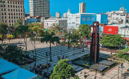 Imagem: Fotografia. Uma praça com bancos e árvores no piso quadriculado. No centro há uma torre com um relógio na ponta. Em volta há vários prédios, construções e pessoas andando. Fim da imagem.