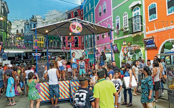 Imagem: Fotografia. No centro há crianças em um coreto. Em volta há várias pessoas e nas laterais, casas coloridas.  Fim da imagem.